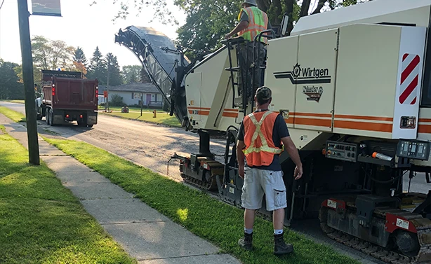 A Peters Paving Wirtgen cold planer milling a residential street and loading reclaimed asphalt into a waiting dump truck via conveyor.