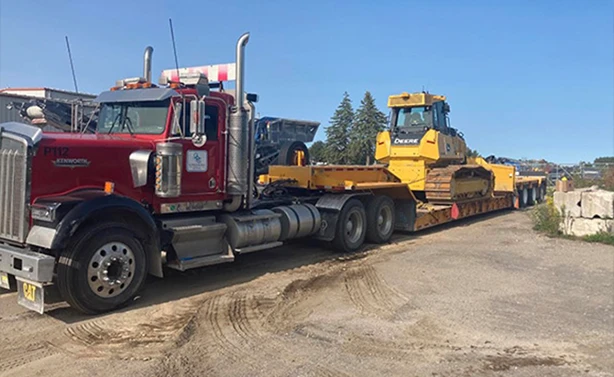 Peters Paving Kenworth semi-truck hauling a John Deere bulldozer on a heavy-duty float trailer to a construction site.