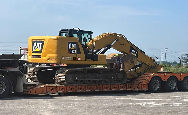 A Peters Paving CAT 323 excavator securely loaded on an orange lowboy trailer for transport to a commercial excavation site.