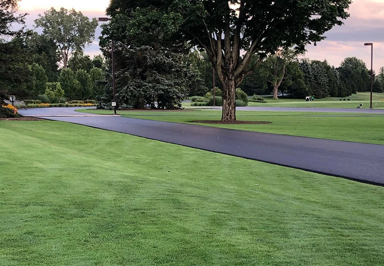 A long, flawless asphalt driveway winding through a professionally maintained golf course and country club property.