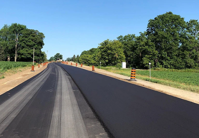 Large scale municipal road paving project featuring a fresh asphalt surface and orange traffic barrels for safety in a rural setting.